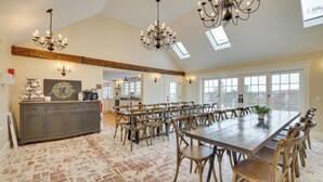 A spacious dining area with a long table and multiple chairs, a kitchen in the background, and a chandelier. - Oceanfront Mid-coast Maine Home w/ Private Beach! (Stockton Springs)