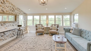 A spacious living room with a light blue sofa, two armchairs, a coffee table, and a patterned rug. A chandelier hangs from the ceiling, and there are large windows providing natural light. - The Beachhouse Retreat w/ Prescot Bay Views! (Stockton Springs)