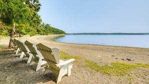 A row of Adirondack chairs on a pebble beach with a calm lake and a forested area in the background. - The Beachhouse Retreat w/ Prescot Bay Views! (Stockton Springs)
