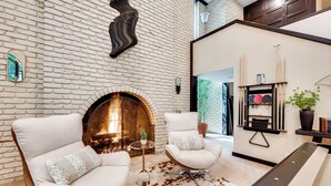 A modern living room with a brick fireplace, two white armchairs, a round coffee table, and a minimalist decor style. - Heated Pool: Tropical Townhome in Wheat Ridge (Wheat Ridge)