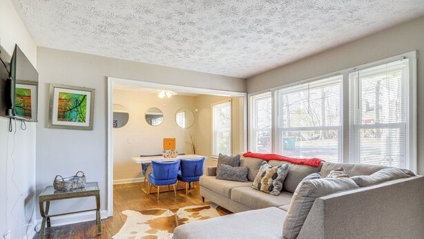 A living room with a grey sofa, a glass coffee table, and a cowhide rug. There are two blue chairs, a small table with a basket, and a TV mounted on the wall. - Pet-friendly Georgia Retreat w/ Fire Pit & Porch! (Forest Park)
