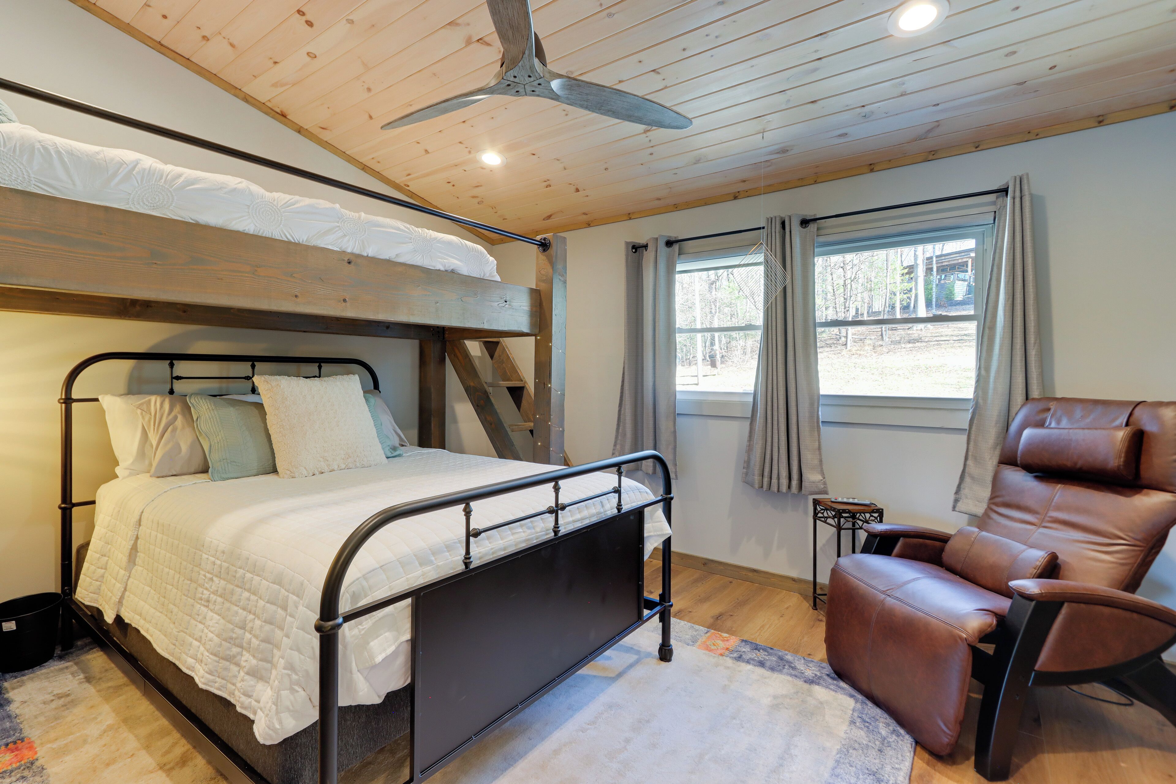Una cama litera con cabecera, una butaca de cuero marrón y un ventilador de techo en una habitación con pisos de madera y una ventana.