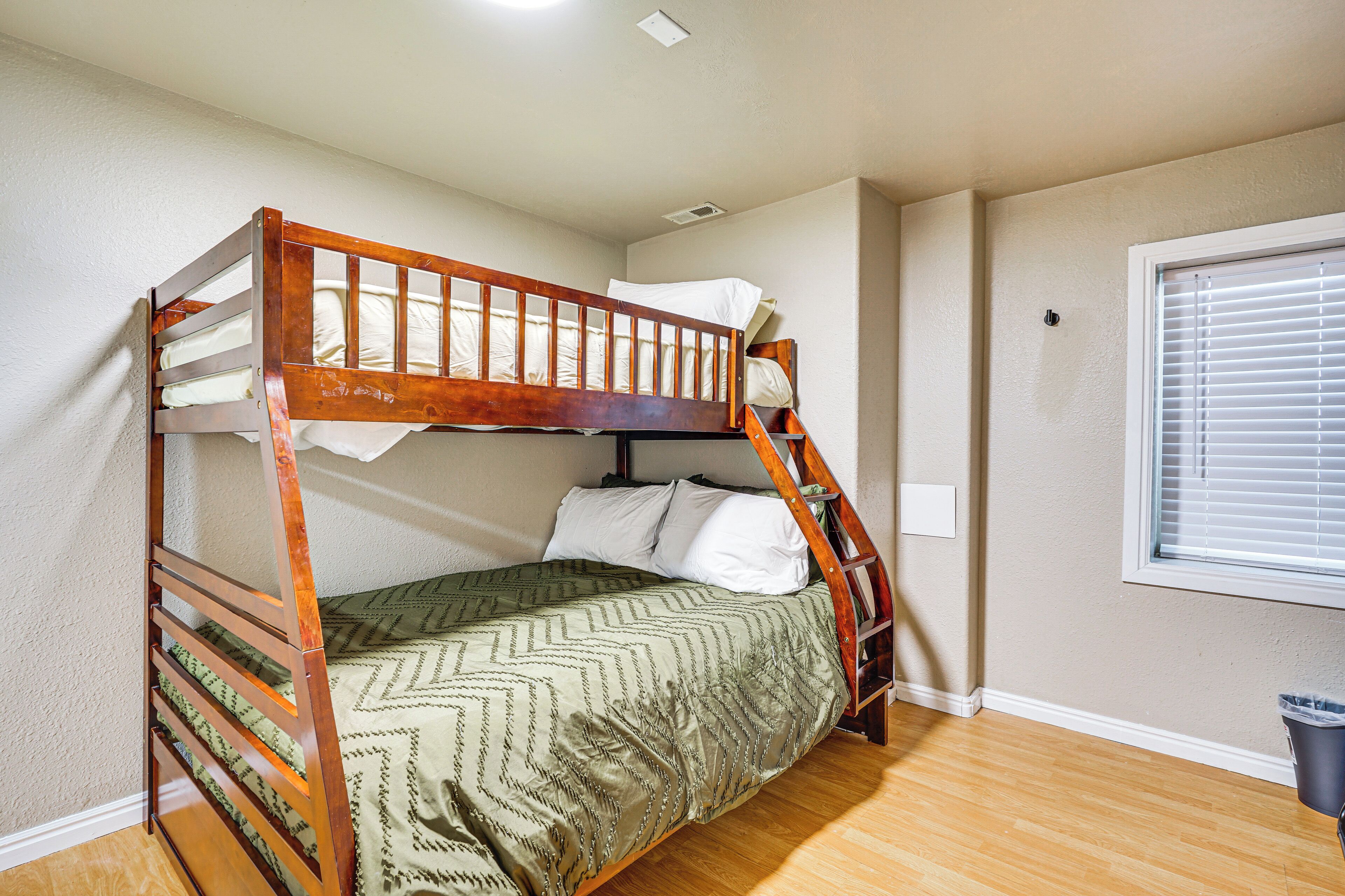 A bunk bed with a ladder, a window with blinds, and a trash can.