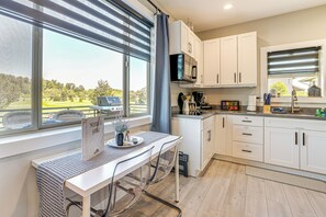 A modern kitchen with white cabinets, a dining table with chairs, and a view of a green landscape through large windows. - Modern Studio Suite in Sandy Point on Lummi Bay! (Ferndale)