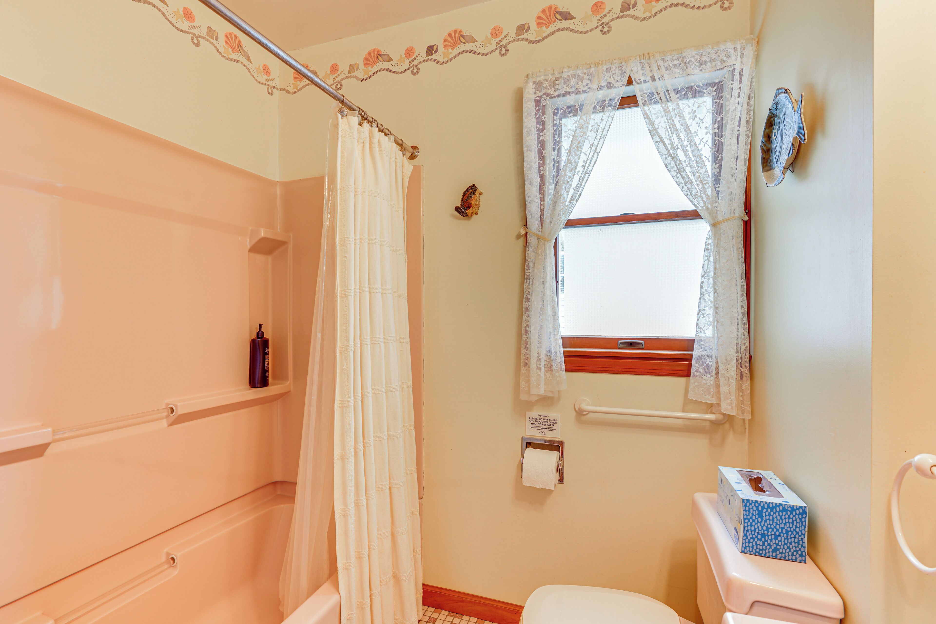 Une salle de bain avec un rideau de douche, des toilettes, une fenêtre ornée de rideaux en dentelle et une étagère contenant des articles de toilette.