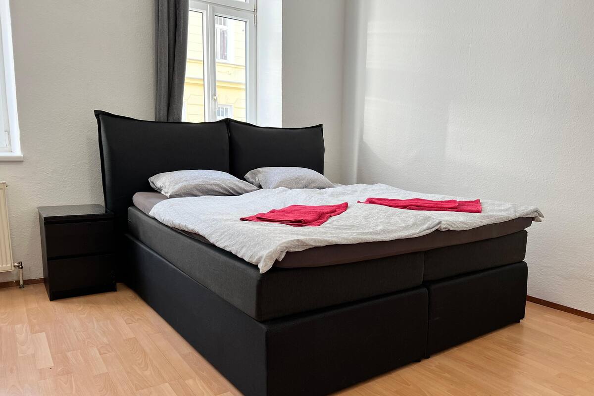 A modern bed with a black frame and grey bedding, a small bedside table, and a window with curtains.