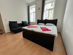 A modern bedroom with a large bed, a sofa, and a view of buildings through the window. - Affordable 3BR shared Apt. near Meidling (Vienna)