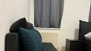 Un petit canapé de couleur sombre, agrémenté d’un seul coussin bleu, est placé contre un mur blanc. Une fenêtre aux rideaux gris et un radiateur se trouvent en dessous.
