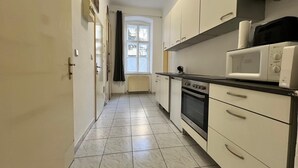 Une cuisine étroite avec des meubles blancs, un four à micro-ondes, un four traditionnel et une fenêtre avec des rideaux.