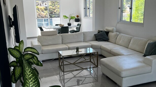 Una sala de estar moderna con un sofá blanco, una mesa de centro de vidrio y un amplio ventanal con vistas al verdor exterior.