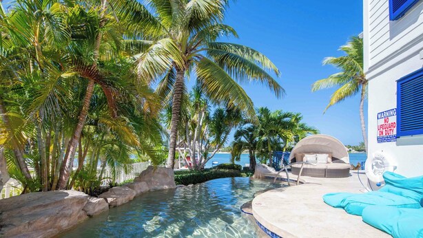 Ein Poolbereich mit klarem Wasser, Palmen und einer Lounge mit blauen Kissen.