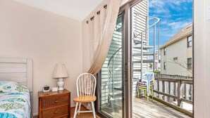 Een slaapkamer met een bed, nachtkastje, stoel, lamp en een schuifdeur naar een balkon met een houten terras en een wenteltrap.