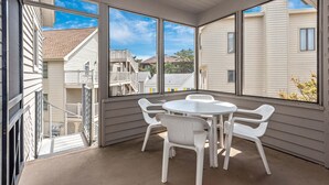 Een overdekt terras met een ronde tafel en witte plastic stoelen, grote ramen met uitzicht op omliggende huizen.