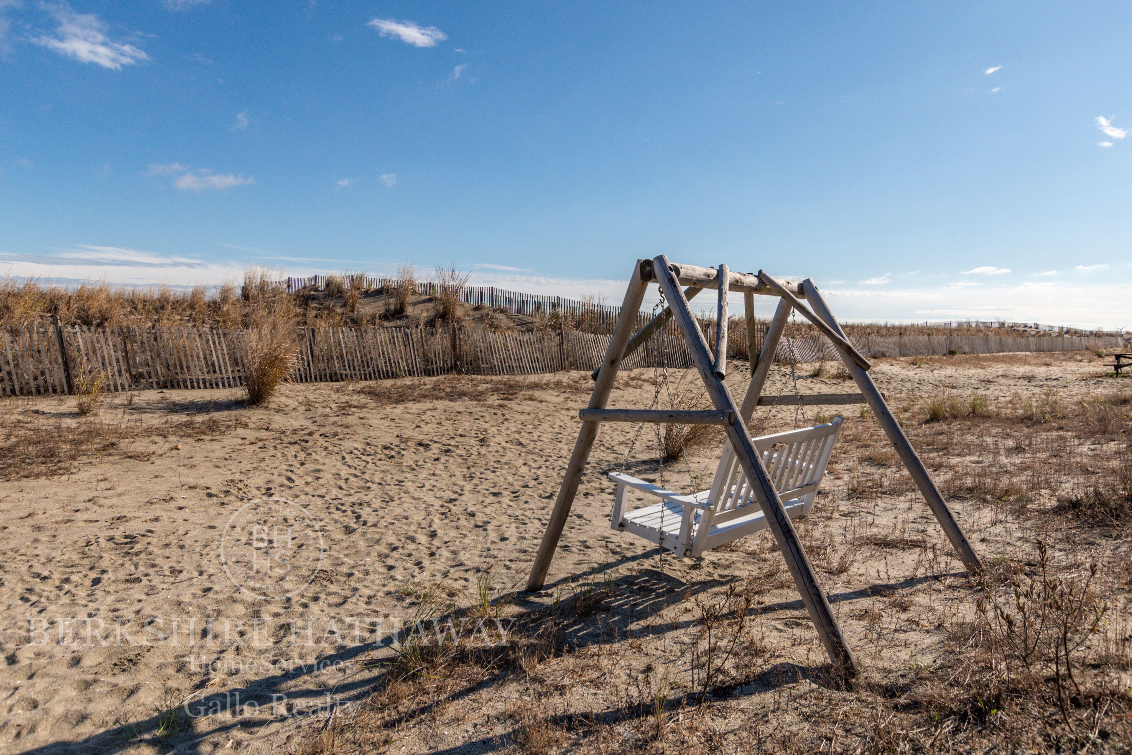 A wooden swing set in a sandy area with a fence and sparse vegetation in the background.