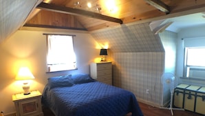 A bedroom with a sloped ceiling, a bed with a blue quilt, a bedside lamp, a dresser, and a window with curtains.