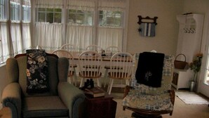 A cozy living room with a sofa, armchair, and ottoman. There is a dining table with chairs and a small table with books and a lamp. A window with curtains and a wall-mounted shelf with a basket are also visible. - Just a block to the beach!  701 Cedar (Lewes)