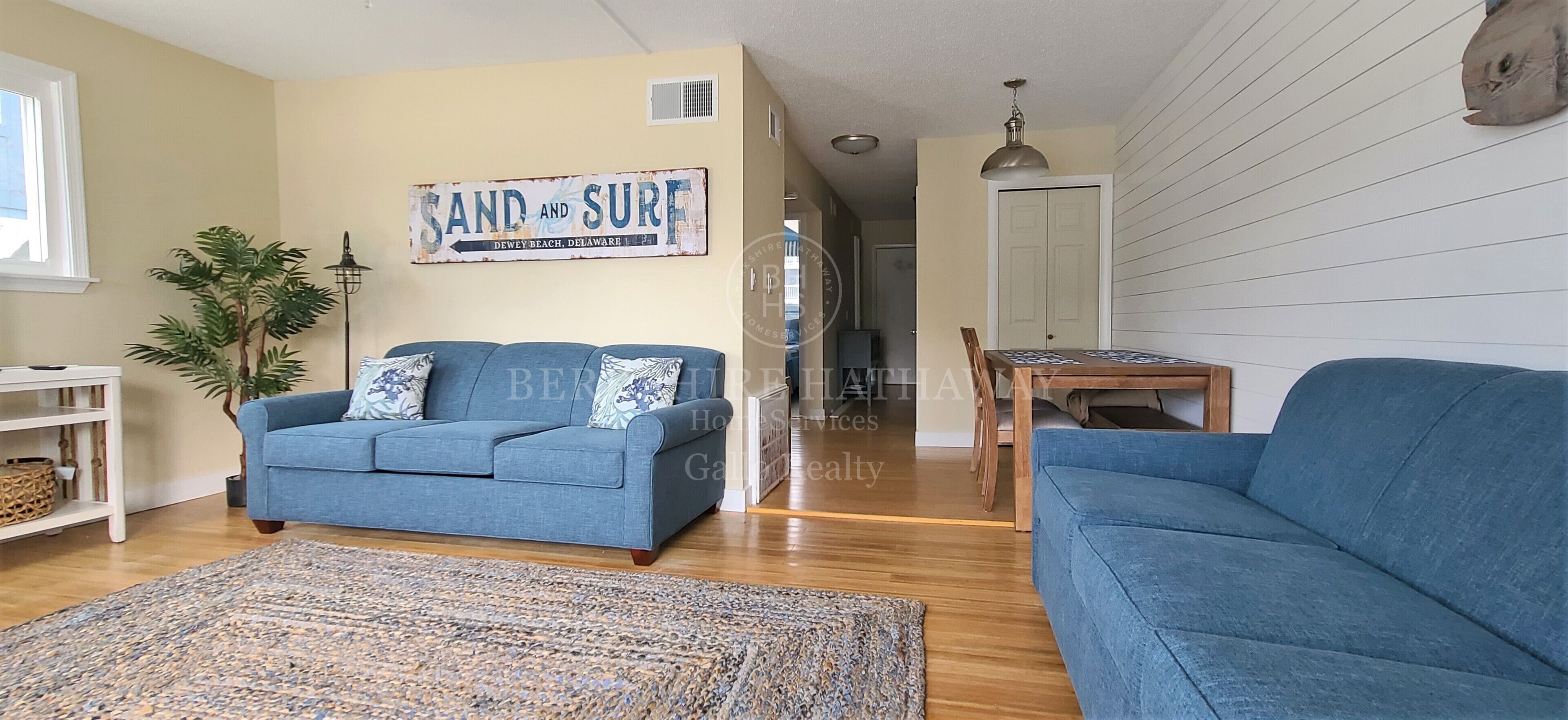 Ett vardagsrum med en blå soffa, ett soffbord och en matta. På väggen hänger en skylt med texten ”SAND OCH SURF”, och i hörnet står en växt.