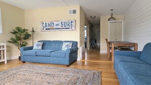 A living room with a blue sofa, a coffee table, and a rug. There is a sign on the wall that reads "SAND AND SURF" and a plant in the corner. - 24 Dickinson Avenue, 8A Dewey Villas, Dewey Beach, Delaware (Dewey Beach)