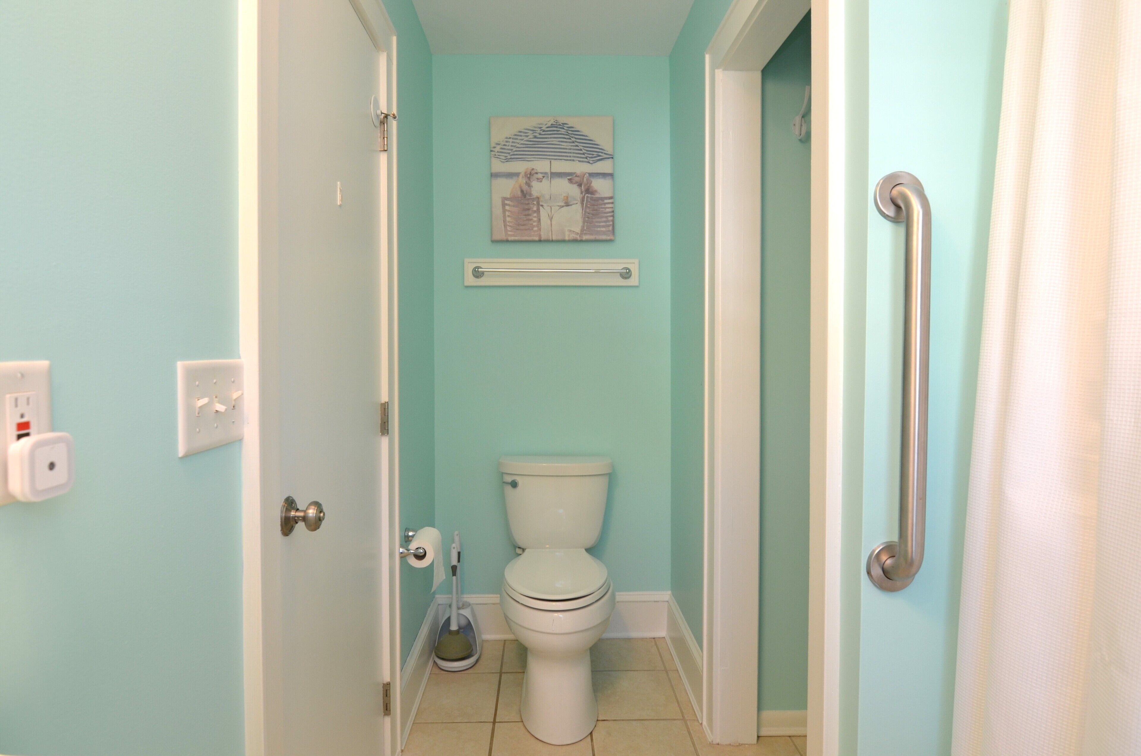 A bathroom with a toilet, a handrail, a painting on the wall, and a floor lamp.