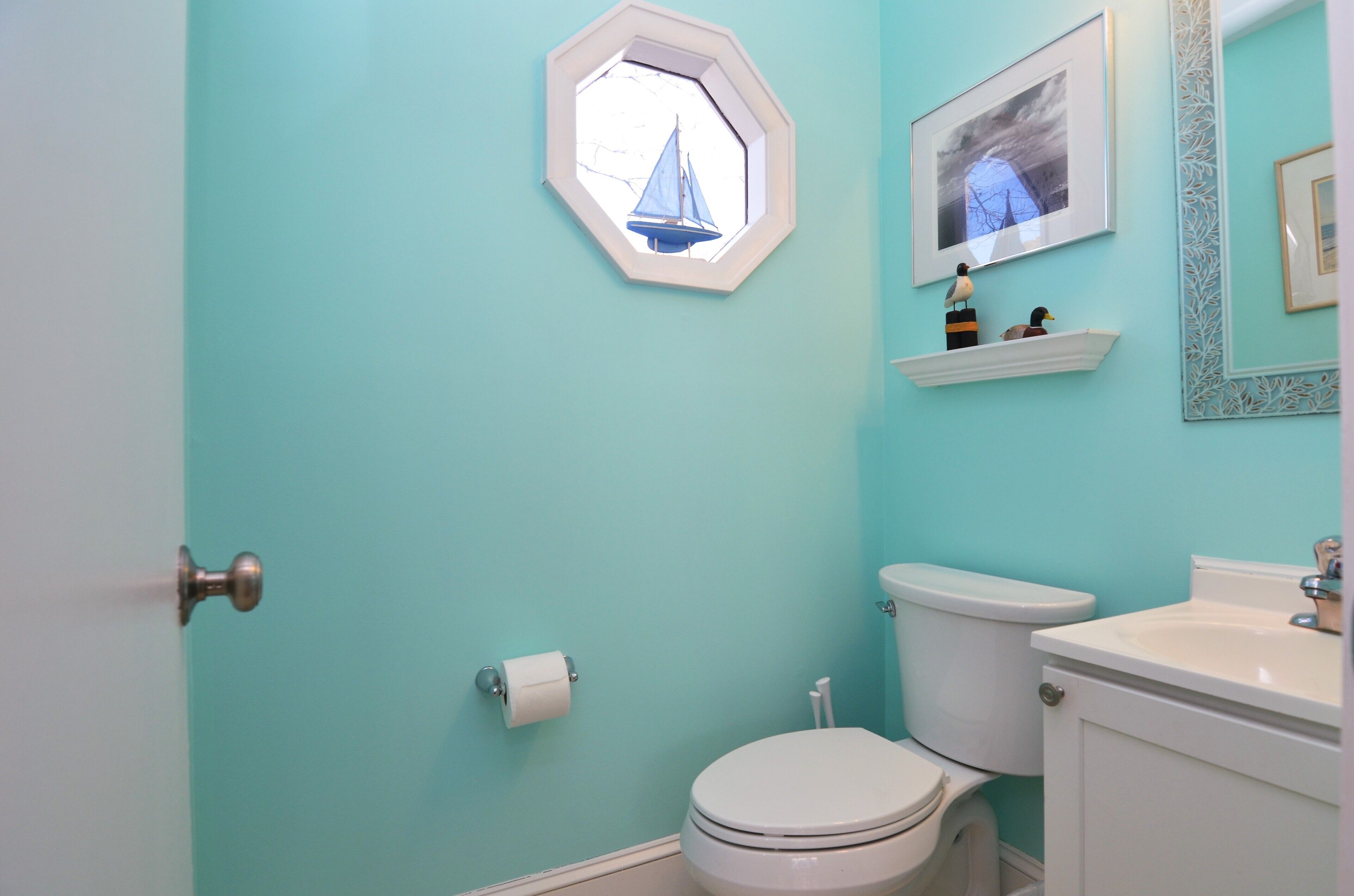 Ein Badezimmer mit hellblauer Wand, einer weißen Toilette, einem weißen Waschbecken, einer weißen Tür und einem gerahmten Bild an der Wand.