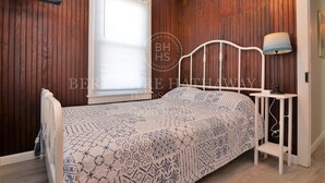 Quarto com parede de painel de madeira, estrutura de cama de metal branca, colcha estampada, mesa de cabeceira com abajur e janela com persianas.