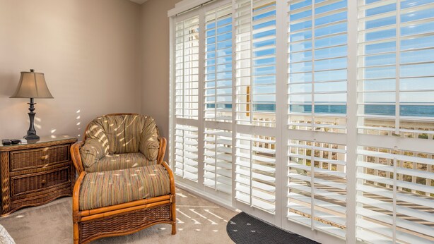 A room with a wicker chair, a lamp, and a view through white blinds to the outdoors.
