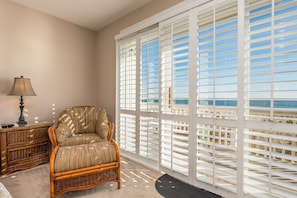 A room with a wicker chair, a lamp, and a view through white blinds to the outdoors.