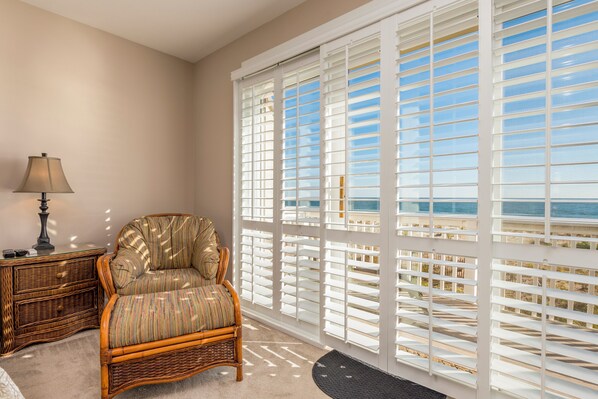 A room with a wicker chair, a lamp, and a view through white blinds to the outdoors.