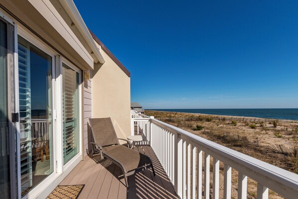 A balcony with two chairs and a table, overlooking a beach with dunes and the ocean.