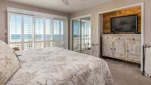 A bedroom with a large bed, a ceiling fan, a TV mounted on a wooden cabinet, and a view of the ocean through large windows.
