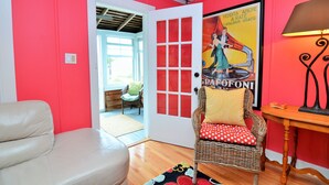 A room with a red wall, a wicker chair with a red cushion, a white leather sofa, a wooden table, and a poster on the wall. - 8 Rhode Island - BAY BLOCK HOME, STEPS FROM LEWES BEACH (Lewes)