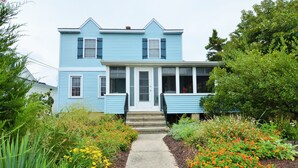 A two-story blue house with a front porch, a concrete pathway leading to the entrance, and a garden with various plants and flowers. - 8 Rhode Island - BAY BLOCK HOME, STEPS FROM LEWES BEACH (Lewes)