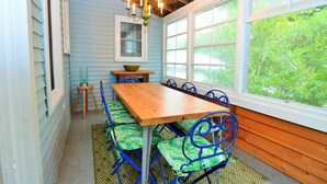 A dining area with a wooden table and blue chairs, a chandelier, and a view of greenery through large windows. - 8 Rhode Island - BAY BLOCK HOME, STEPS FROM LEWES BEACH (Lewes)