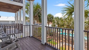 A covered outdoor area with a wooden deck, white railings, and a view of a swimming pool and palm trees. - Lazy Beach, Spacious + Shared Pool & 5-Min Walk to the Sand! (Panama City Beach)