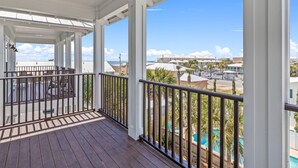 A covered outdoor area with a wooden deck, white railings, and a view of a swimming pool and buildings in the background. - Lazy Beach, Spacious + Shared Pool & 5-Min Walk to the Sand! (Panama City Beach)