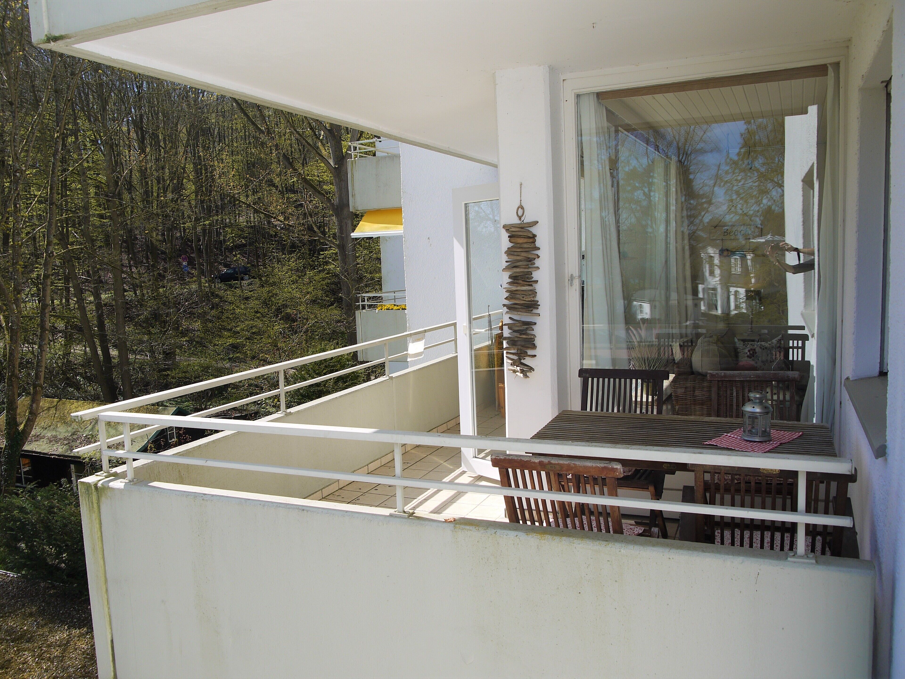 En balkon med et bord og stole, en hængende plante samt udsigt til træer.