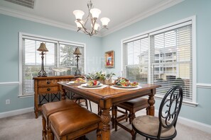 Une salle à manger avec une table en bois dressée pour quatre personnes, flanquée de deux fenêtres équipées de stores, d’un lustre et d’un buffet sur lequel est posée une lampe.