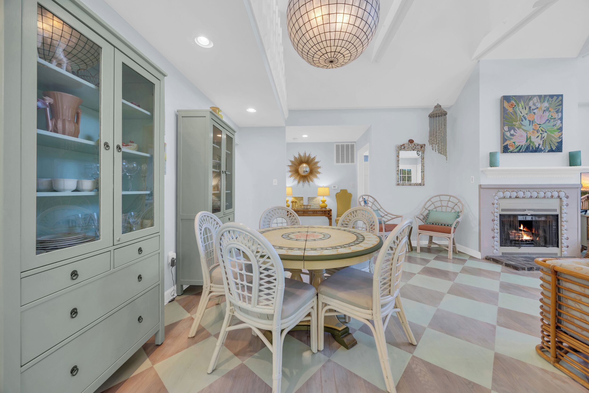 A dining area with a round table, white chairs, a cabinet with glass doors, a chandelier, a fireplace, and a checkered floor.