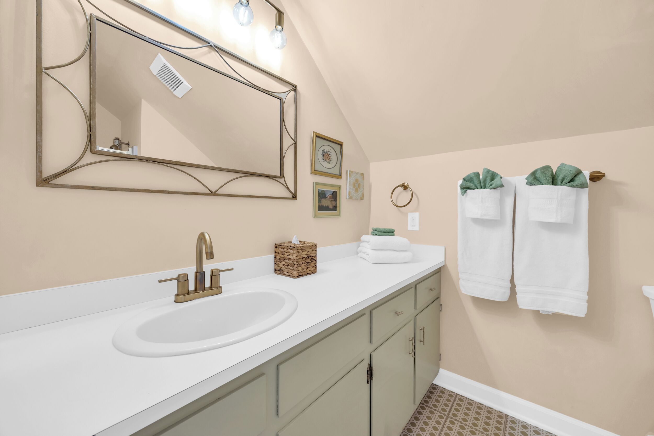 A bathroom with a white sink, gold faucet, and a large mirror with a decorative frame. There are towels and a small basket on the counter.