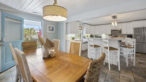 A dining area with a wooden table and chairs, a hanging light fixture, and a view of the kitchen with white cabinets and stainless steel appliances. - Peace of Pawleys - Creek Front, 3-bedroom (Pawleys Island)
