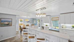 A modern kitchen with white cabinetry, a bar area with stools, and a dining area with a view of the outdoors. - Peace of Pawleys - Creek Front, 3-bedroom (Pawleys Island)