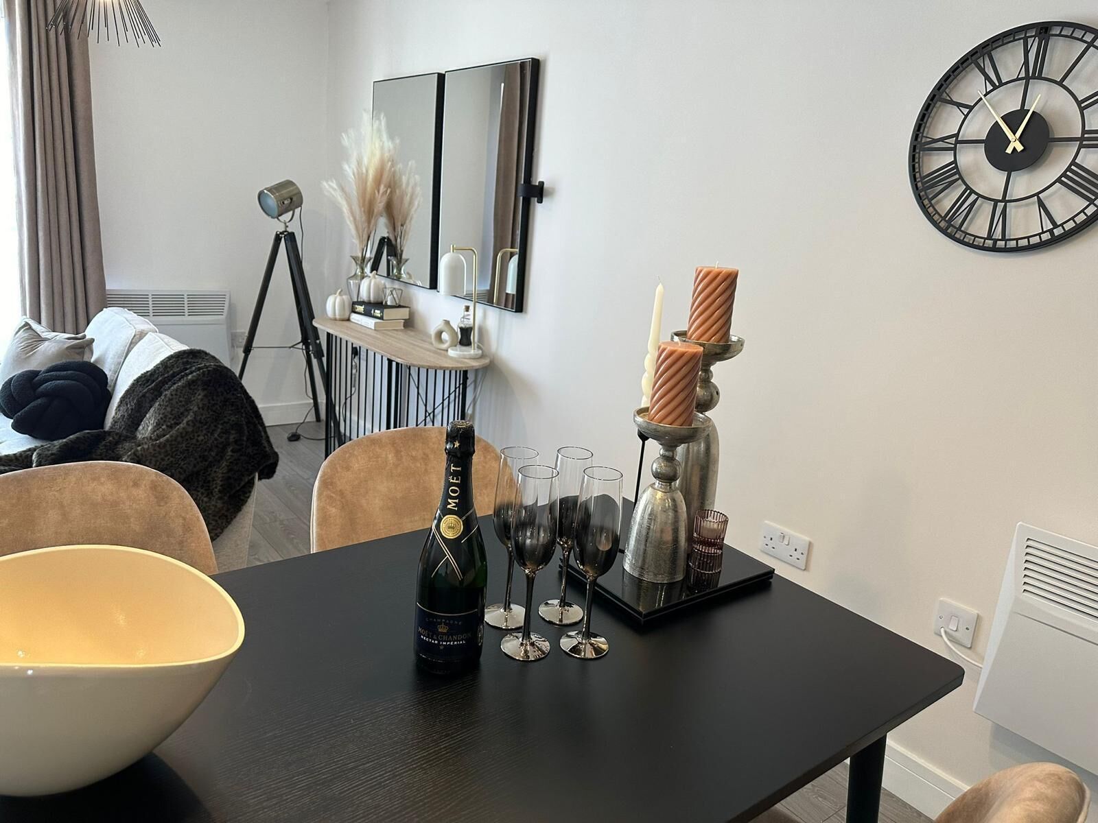 A modern living room with a dark wooden table set for a meal, a large wall clock, and a mirror reflecting the room.