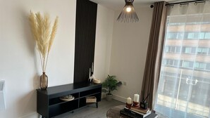 Un salon moderne avec un parquet en bois foncé, un meuble de télévision noir, une table basse ronde en verre et une grande fenêtre ornée de rideaux légers.