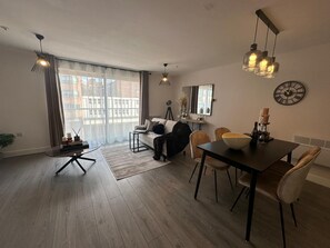 A modern living room with a sofa, dining table, and chairs. There is a large window with curtains, a floor lamp, and a wall clock. - The Nest 1-bed Apartment in Hemel Hempstead (Hemel Hempstead)