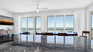 Amplia habitación con un gran comedor, varias ventanas con vistas al océano, un ventilador de techo y suelo de mármol.