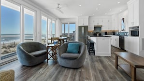 Una cocina moderna con armarios blancos, una isla grande y vistas al océano. Hay dos sillones grises y una mesa de comedor de madera.