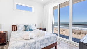 Un dormitorio con una cama grande, una mesita de noche, una lámpara y vistas a la playa a través de amplias ventanas.