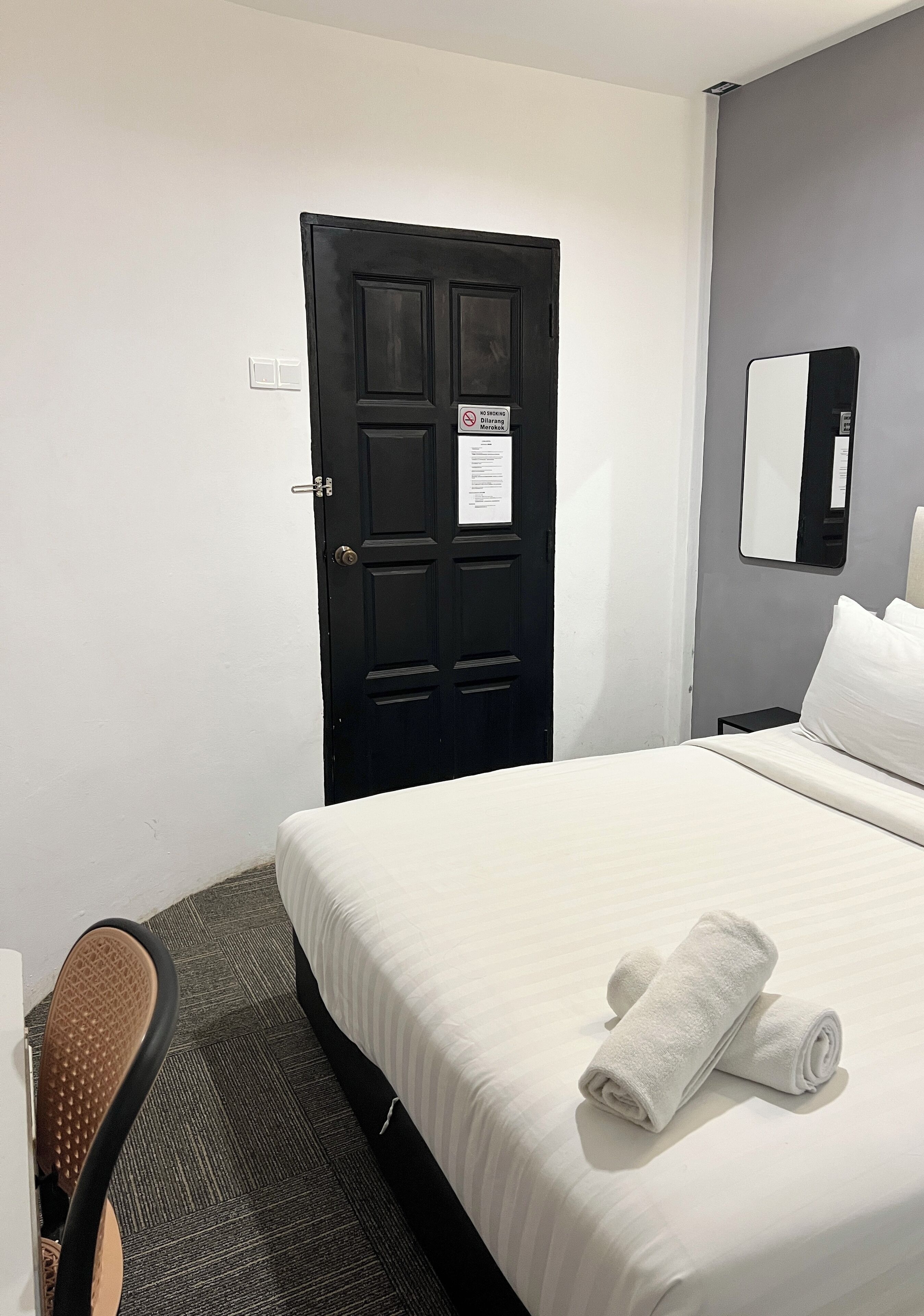 A hotel room with a bed, a chair, a black door, and a mirror on the wall.