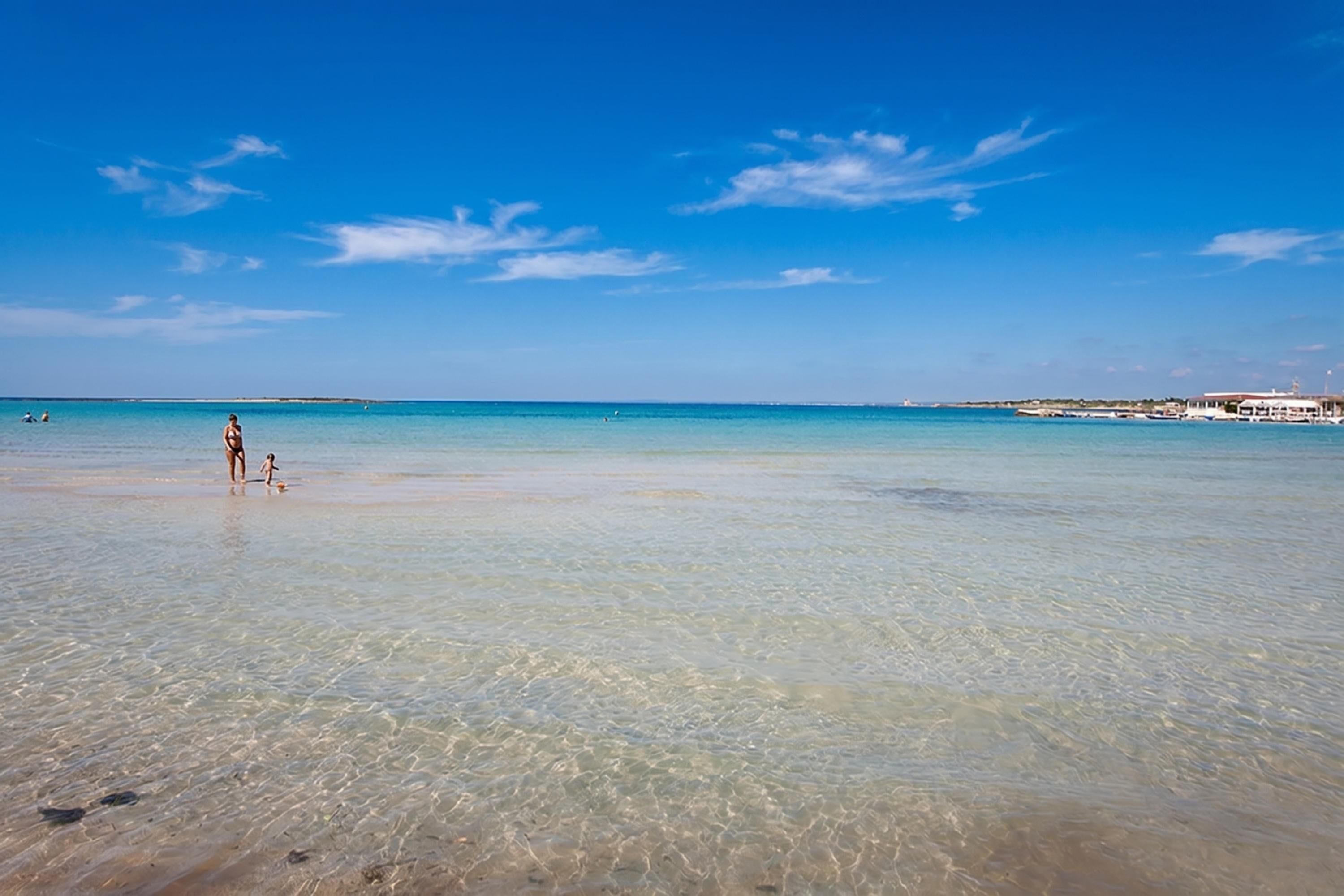 Una playa con agua cristalina, una persona de pie y un edificio a lo lejos.