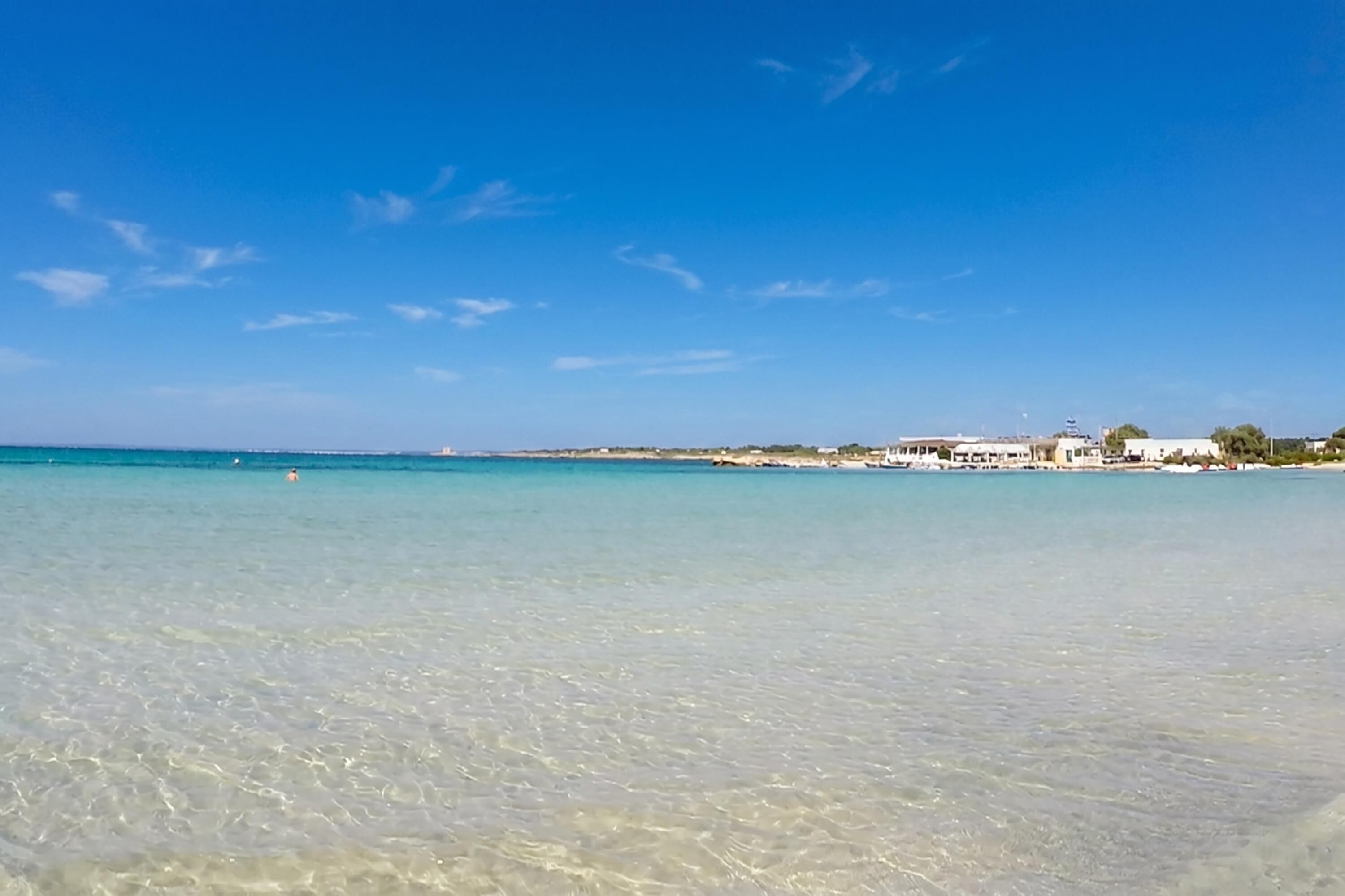 Escena de playa costera con agua cristalina, algunos edificios y una línea de costa distante.